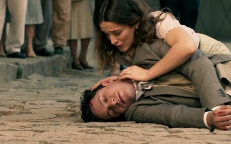 O ator Rainer Cadete, o Celso, com um ferimento na cabeça está caído em uma rua de paralelepípedos, enquanto Bianca Bin, a Maria, o socorre em cena de Êta Mundo Bom!