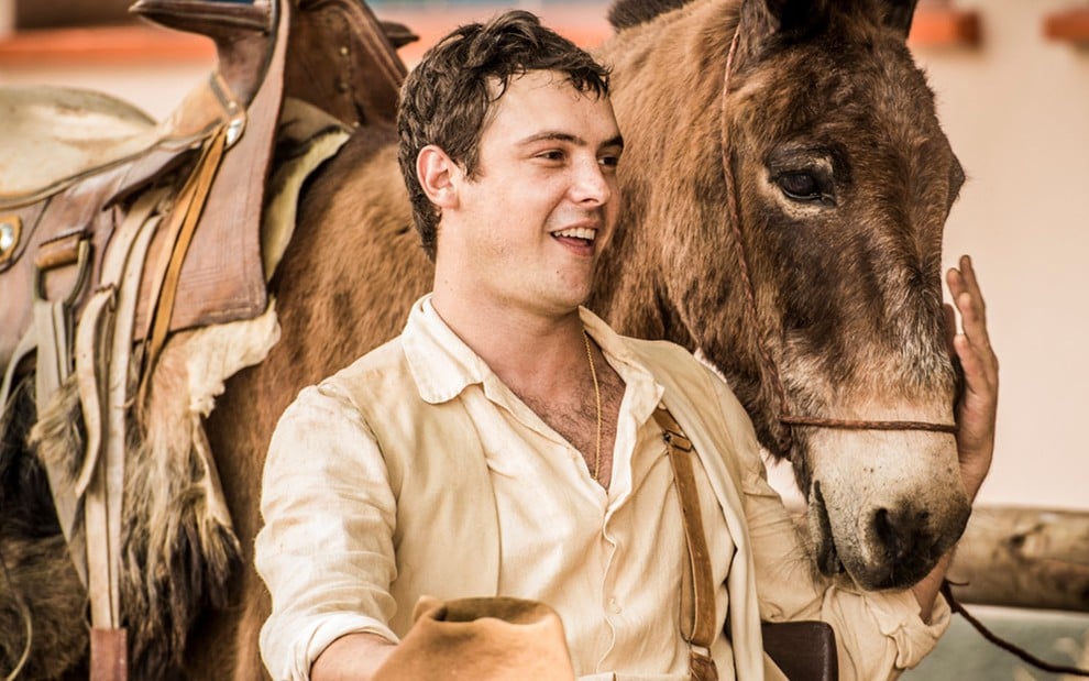 O ator Sergio Guizé caracterizado como Candinho sentado ao lado de um burro em cena de Eta Mundo Bom