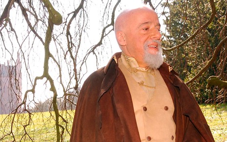 O escritor Paulo Coelho com uma casaca vermelha e barbicha branca caracterizado como o mago Simon em cena de Eterna Magia
