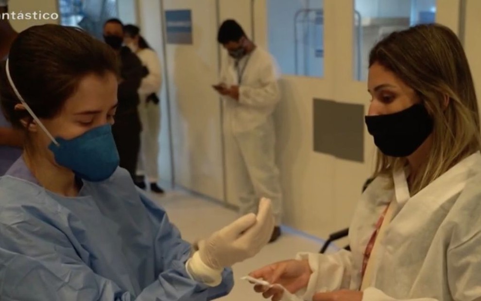 A atriz Marjorie Estiano de máscara e ajeitando luva cirúrgica como a personagem Carolina da série Sob Pressão, da Globo, enquanto é auxiliada por uma assistente 