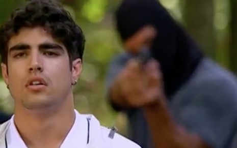 O ator Caio Castro com expressão perplexa caracterizado como o Antenor com um bandido com uma touca preta cobrindo o rosto e revolver apontado para ele às suas costas em cena de Fina Estampa