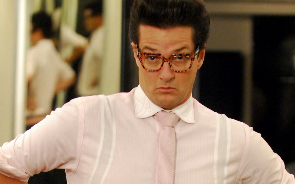 O ator Marcelo Serrado caracterizado como Crodoaldo Valério isa óculos na cor vermelha e gravata em tom rosa em cena de Fina Estampa