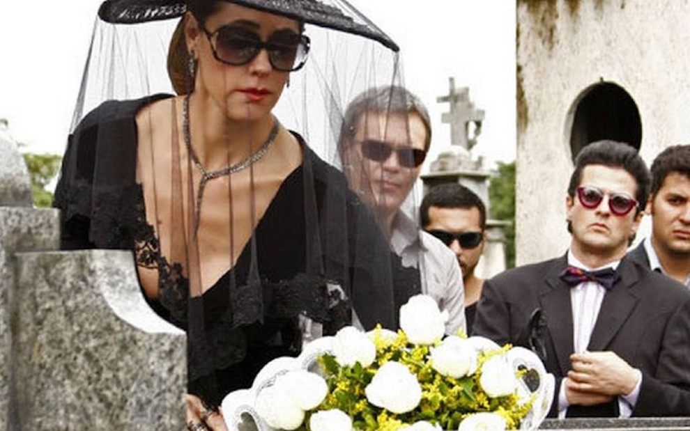 Christiane Torloni usa chapéu preto e véu durante cena de enterro em que coloca flores em um túmulo em Fina Estampa