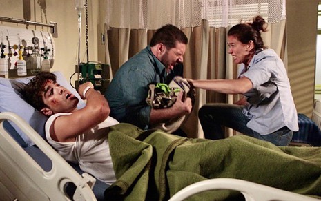 Caio Castro deitado em um um leito de hospital demonstra falta de ar enquanto Lilia Cabral (à direita) simula luta com o intérprete de um bandido