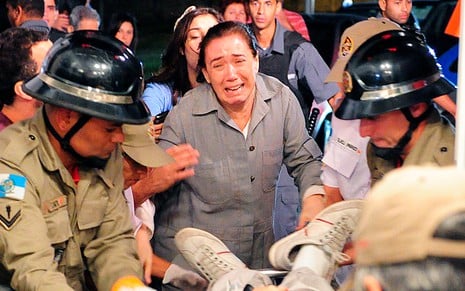 A atriz Lilia Cabral está de macacão cinza, com expressão de choro, sendo segurada por pessoas enquanto um jovem é colocado em uma ambulância em cena de Fina Estampa