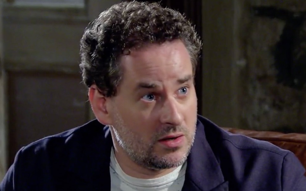 Dan Stulbach caracterizado como Paulo em Fina Estampa: personagem tem expressão de atenção e usa camiseta cinza com blusa azul marinho