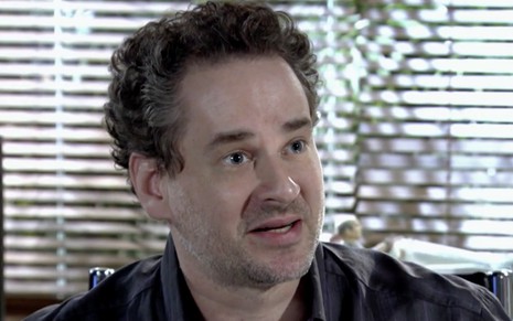 Dan Stulbach caracterizado como Paulo em Fina Estampa; personagem faz cara de contrariado e usa uma camisa social cinza