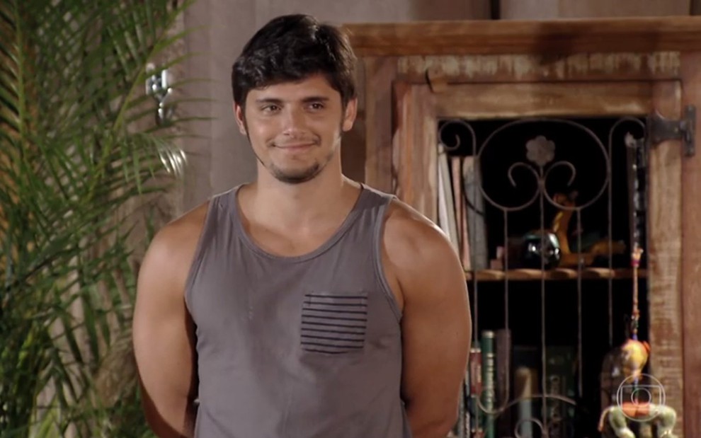 O ator Bruno Gissoni, usando uma camiseta regata branca, com um sorriso no rosto, em cena como Juliano na novela Flor do Caribe