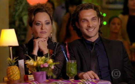 A atriz Claudia Netto e o ator Igor Rickli, sentados em uma mesa de bar, como Guiomar e Alberto, em cena de Flor do Caribe