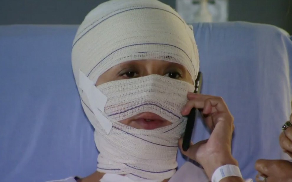 A atriz Cláudia Netto fala em um celular com o rosto completamente coberto por bandagens em cena de Flor do Caribe