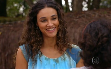 A atriz Débora Nascimento, com sorriso no rosto, usando um vestido azul, em cena como Taís em Flor do Caribe