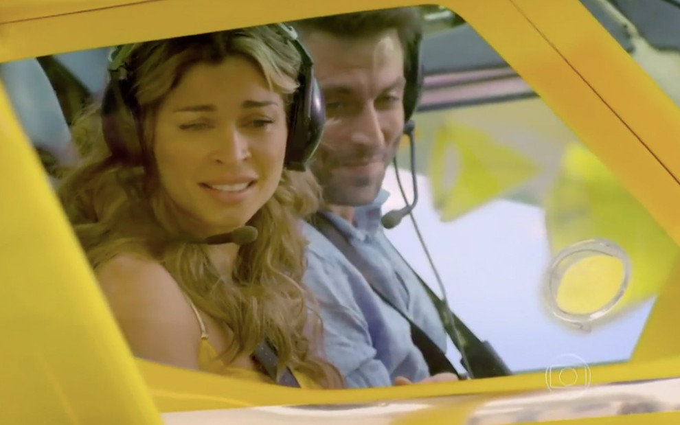 Dentro de um avião amarelo, a atriz Grazi Massafera como Ester e o ator Henri Castelli como Cassiano olham emocionados para terra firme em cena de Flor do Caribe