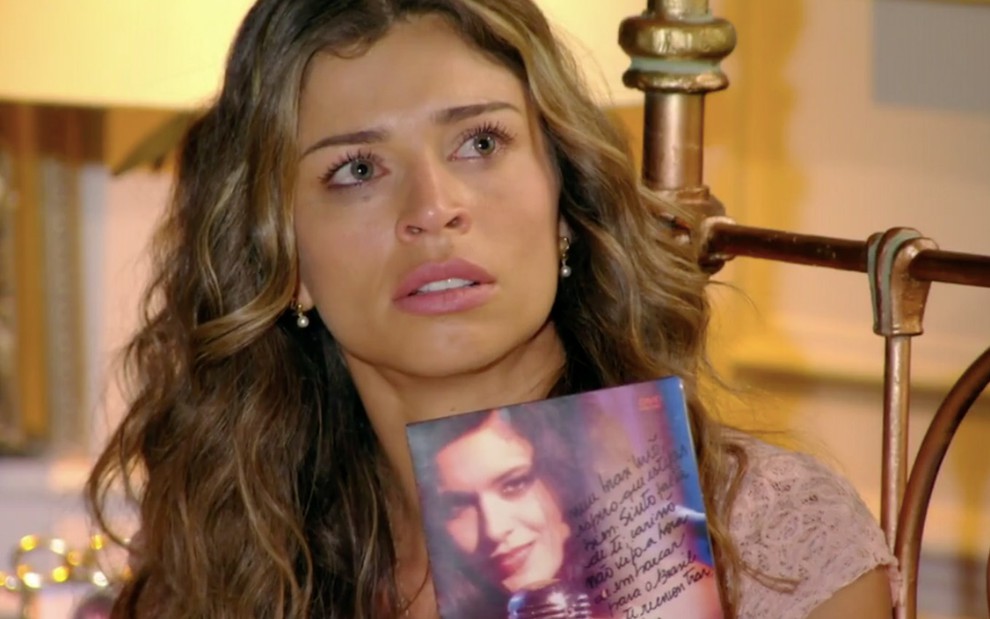 A atriz Grazi Massafera como Ester com a maquiagem borrada por chorar diante de um DVD com a imagem de Moro Anghileri como Cristal em cena de Flor do Caribe