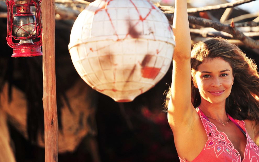 A atriz Grazi Massafera segura em uma armação de madeira com braços levantados em uma praia caracterizada como a Ester de Flor do Caribe