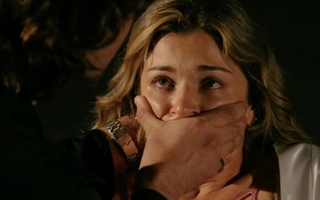 A atriz Grazi Massafera como Ester está caida no chão com lágrimas nos olhos e uma mão tapando sua boca com violência em cena de Flor do Caribe
