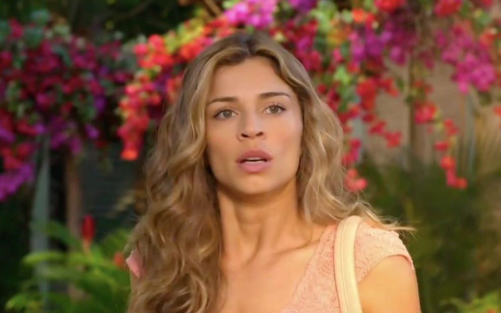 A atriz Grazi Massafera com expressão de surpresa diante de um muro com várias flores caracterizada como a Ester em cena de Flor do Caribe