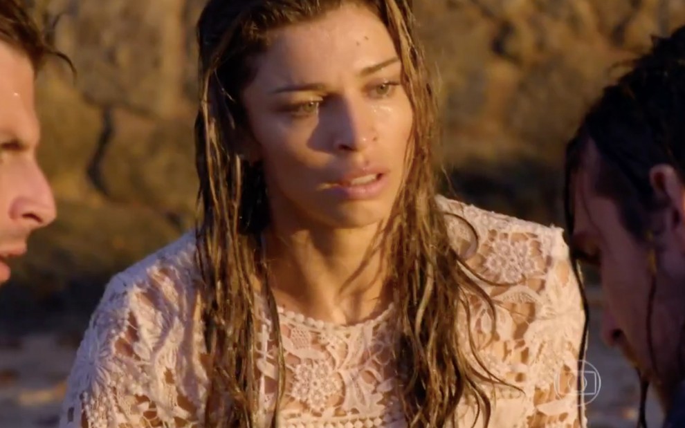 A atriz Grazi Massafera vestida de noiva, completamente molhada, está sentada em uma praia em cena de Flor do Caribe