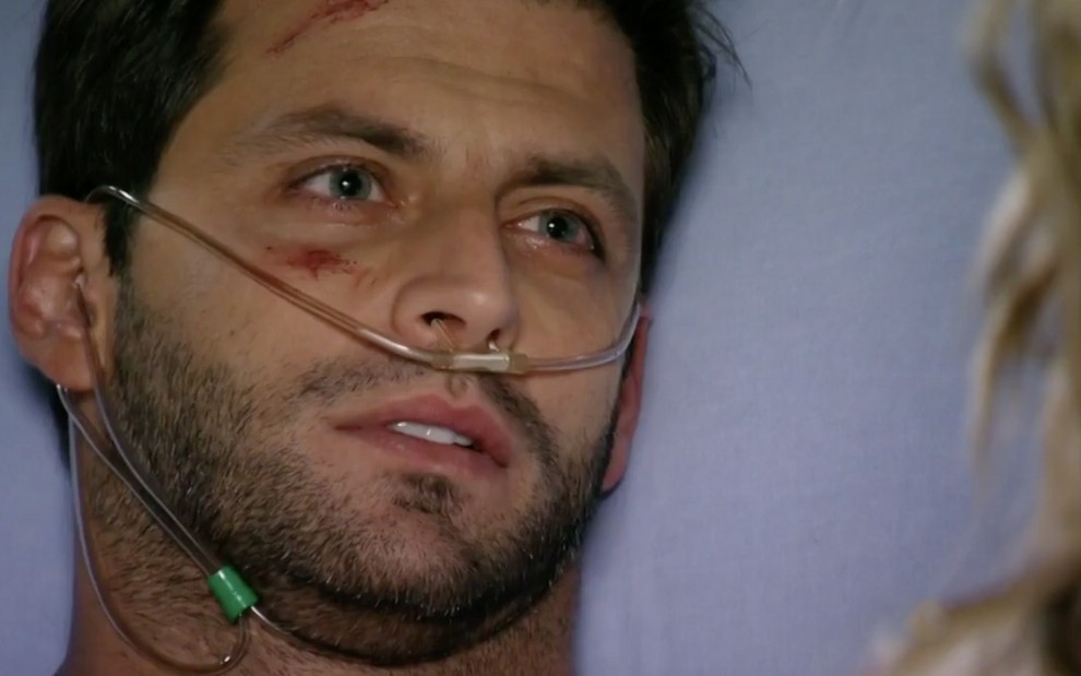 O ator Henri Castelli caracterizado como Cassiano está deitado em um leito de hospital com tubos na frente da nariz em cena de Flor do Caribe