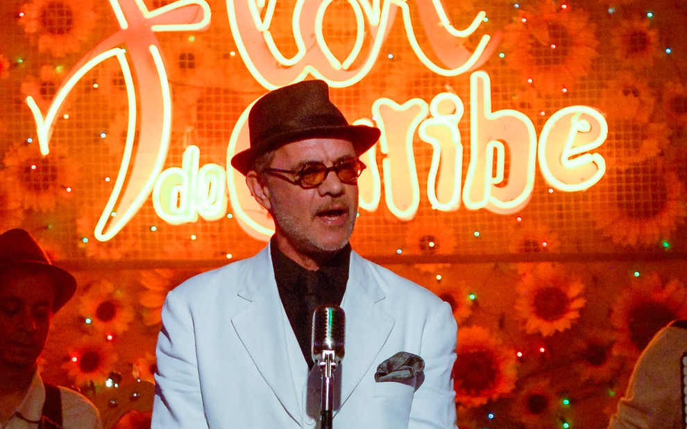O ator Jean Pierre Noher de roupa de gala, chapéu e óculos escuros em frente ao logo da novela Flor do Caribe que ficava em um bar homônimo no folhetim