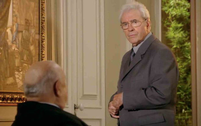 O ator Sérgio Mamberti, de costas, sentado em uma cadeira de rodas como o Dionísio encara Juca de Oliveira, o Samuel, diante de uma reprodução do quadro A Arte da Pintura, de Vemeer