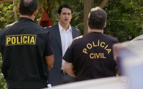 O ator Raphael Vianna com expressão de surpresa como Hélio com dois policias de costas, cujas jaquetas lê-se "polícia civil" em cena de Flor do Caribe