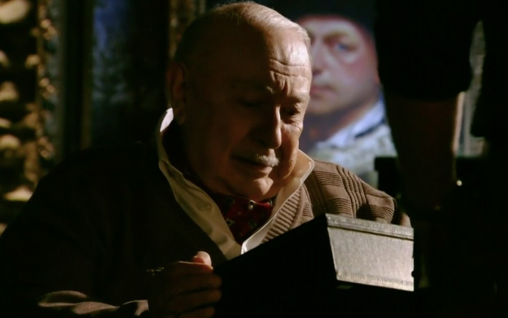 O ator Sérgio Mamberti como Dionísio sentado em um quarto escuro, com uma caixa nas mãos e uma pintura de um homem ao fundo em cena de Flor do Caribe