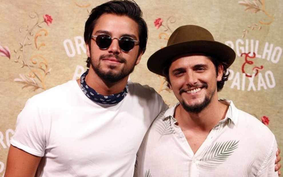 Os atores e irmãos Rodrigo Simas e Bruno Gissoni sorriem na frente de backdrop da novela Orgulho & Paixão