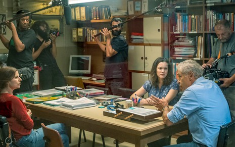Os atores Debora Bloch e Paulo Gorgulho gravam cena da série Segunda Chamada, cercados de técnicos, em uma sala repleta de armários e livros