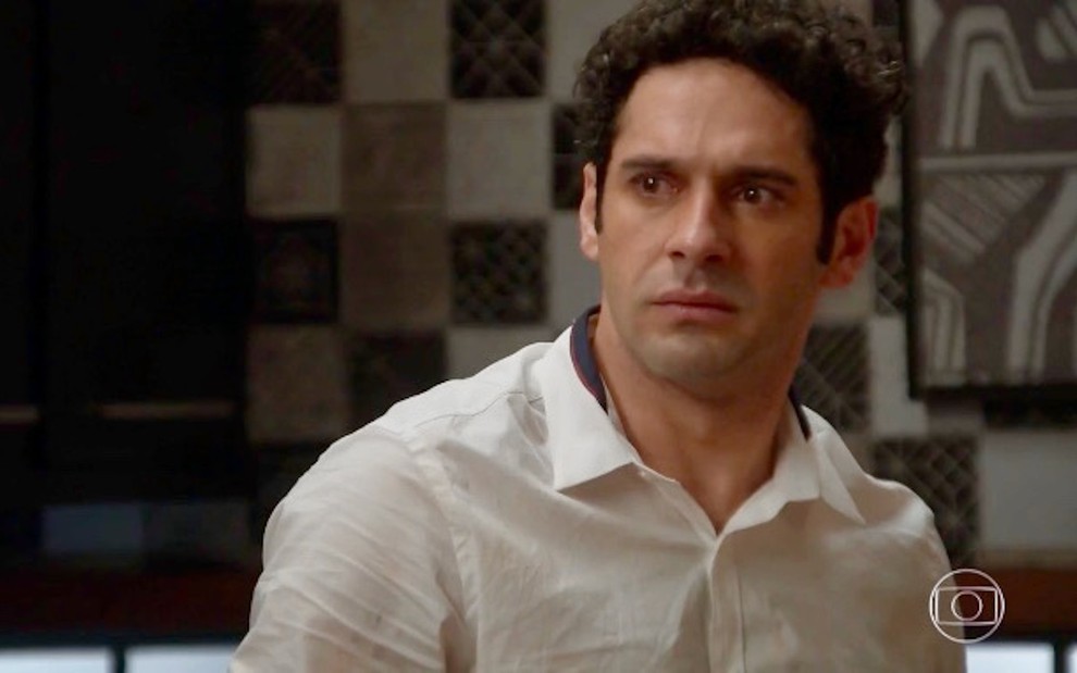 O ator João Baldasserini usa camisa clara e faz uma expressão de revolta em cena de Haja Coração, novela da Globo