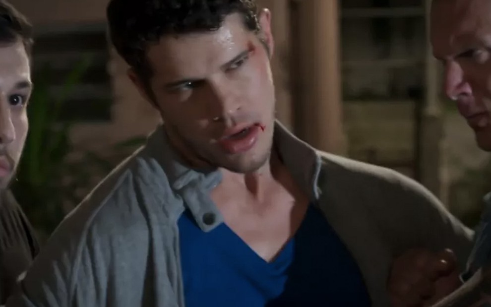 O ator José Loreto como Adônis com o rosto todo machucado é segurado por dois homens em cena de Haja Coração