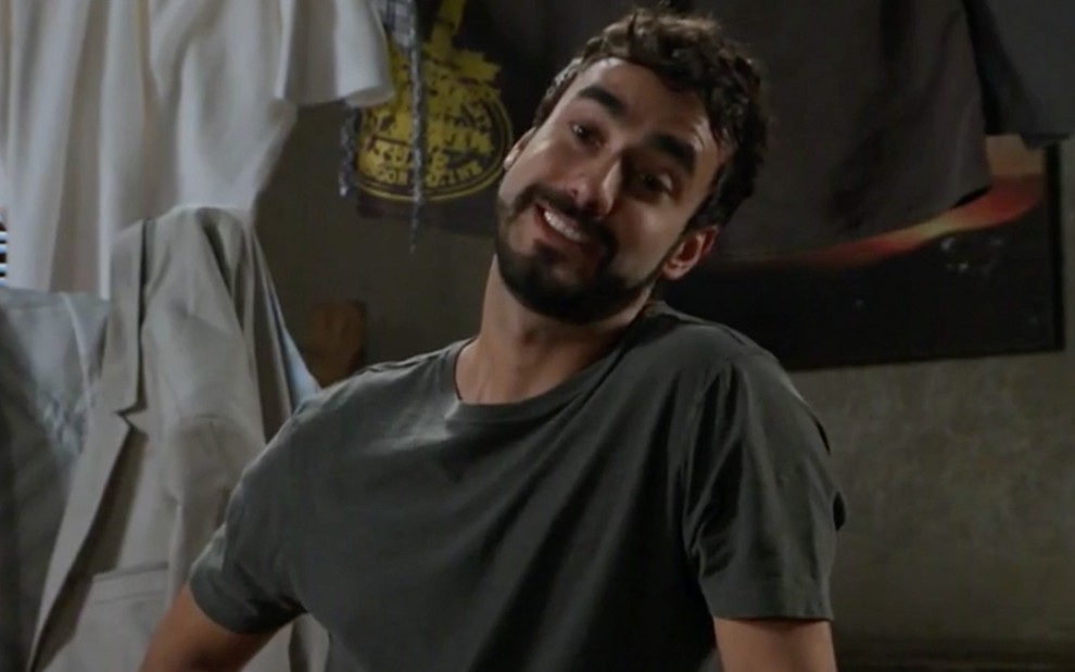 Gabriel Godoy caracterizado em cena de Haja Coração: personagem usa camisa cinza e olha com deboche para alguém fora do quadro