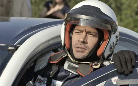 O ator Malvino Salvador abre a porta e sai de um carro de corrida com capacete, luvas e macacão caracterizado como Apolo em cena de Haja Coração