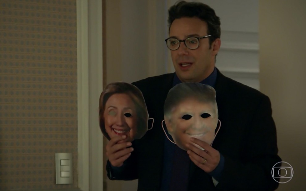O ator Marcelo Médici caracterizado como Gigi segura uma máscara de papel de Hillary Clinton com buraco nos olhos na mão direita e outra de Donald Trump com a mão esquerda