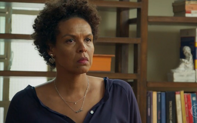 Ana Carbatti grava de camisa azul, colar e brinco prata e expressão tensa como Nair de Haja Coração