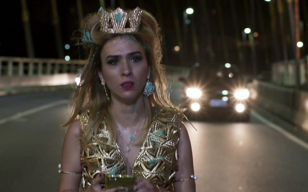 A atriz Tatá Werneck com uma roupa dourada e maquiagem borrada como Fedora Abdala anda em uma ponte entre os carros em cena de Haja Coração