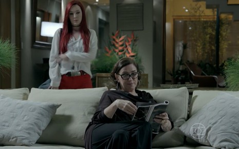 Josie Pessôa grava de cabelo vermelho e roupa branca atrás de Lilia Cabral, sentada no sofá, em cena de Império, novela das nove