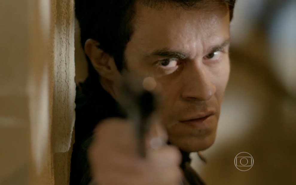 Erom Cordeiro grava escondido, apontando uma arma com olhar sinistro como Fernando de Império, novela das nove da Globo
