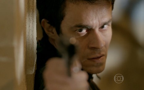Erom Cordeiro grava escondido, apontando uma arma com olhar sinistro como Fernando de Império, novela das nove da Globo