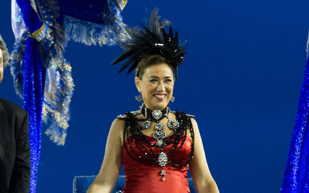 A atriz Lilia Cabral está como Maria Marta com um vestido vermelho e diversos colares de contas pretas, com um fascinator de penas pretas em cena de Império