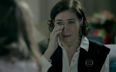 Lilia Cabral grava com camisa branca e colete preto com a mão enxugando uma lágrima como Maria Marta em Império