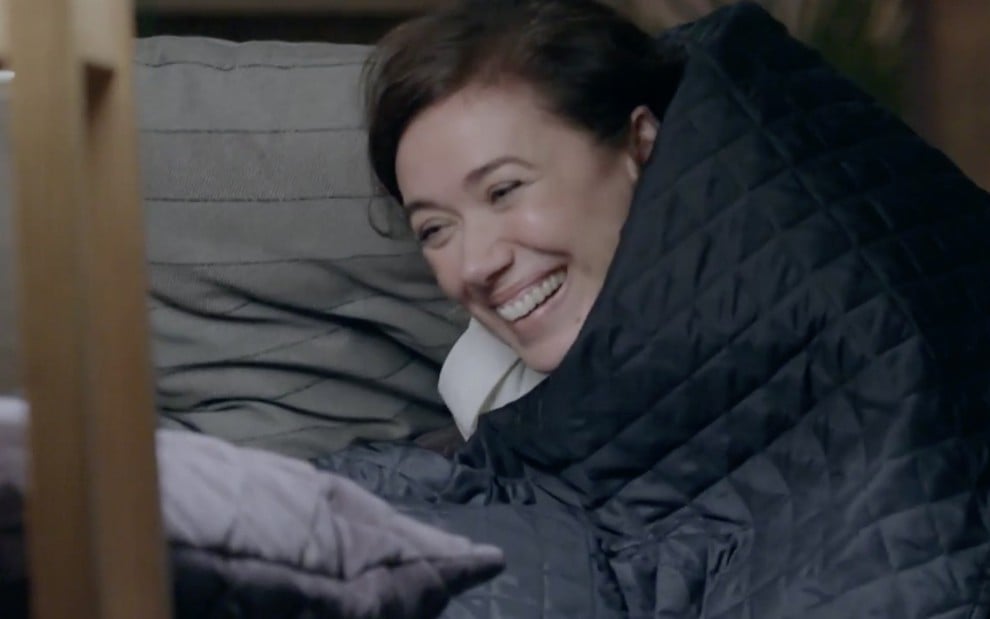 A atriz Lilia Cabral como Maria Marta está deitada em uma cama, aos risos, enrolada em um edredom azul marinho