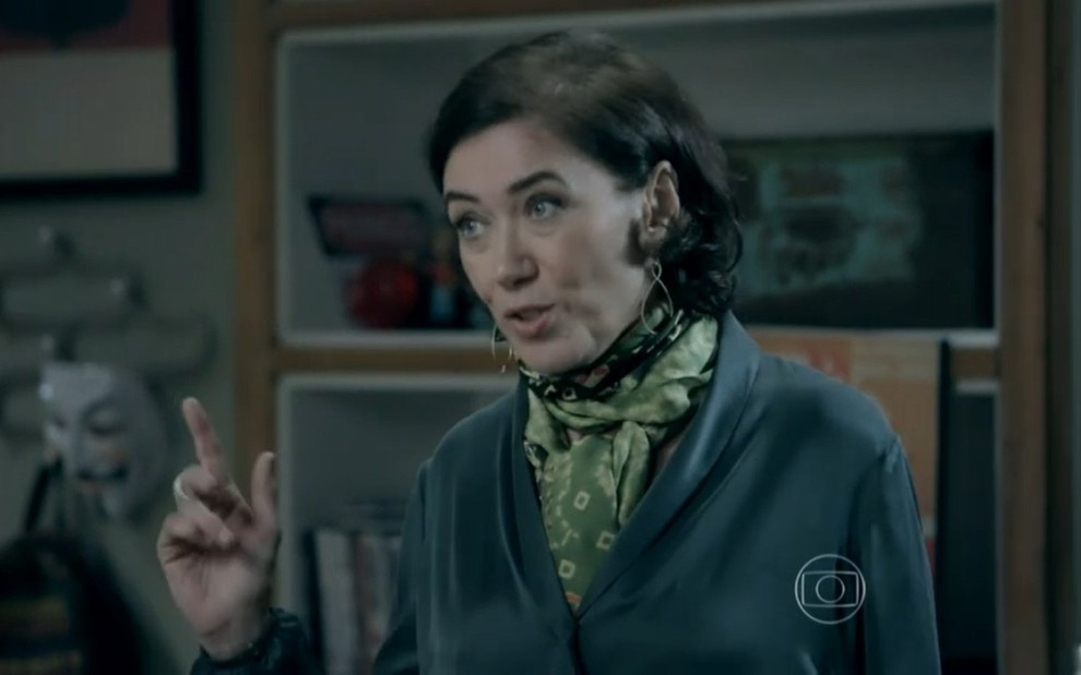 Lilia Cabral grava com camisa azul, lenço verde estampado no pescoço, expressão séria e dedo levantado como Maria Marta