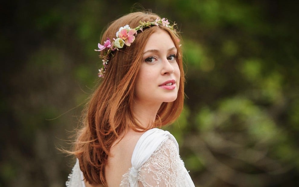 A atriz Marina Ruy Barbosa com uma coroa de flores na cabeça e um vestido branco, ela está em uma floresta, de costas, e vira a cabeça em direção ao fotógrafo como Maria ísis em Império