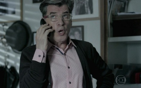 O ator Paulo Betti está em um escritório segurando um celular com a mão direita e faz uma expressão afetada como Téo Pereira em cena de Império