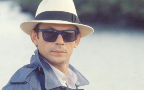O ator José Wilker, de chapéu, óculos escuros e expressão séria, em cena como Roque Santeiro na novela Roque Santeiro