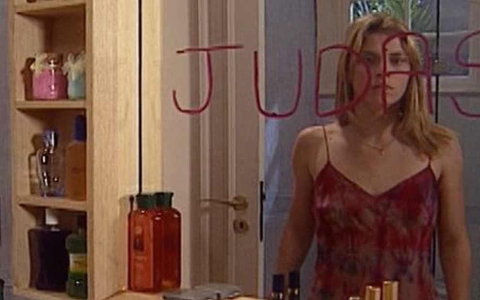 A atriz Carolina Dieckmann se olha no espelho com surpresa caracterizada como Camila, o seu reflexo é tampado pela palavra Judas em batom vermelho em cena de Laços de Família