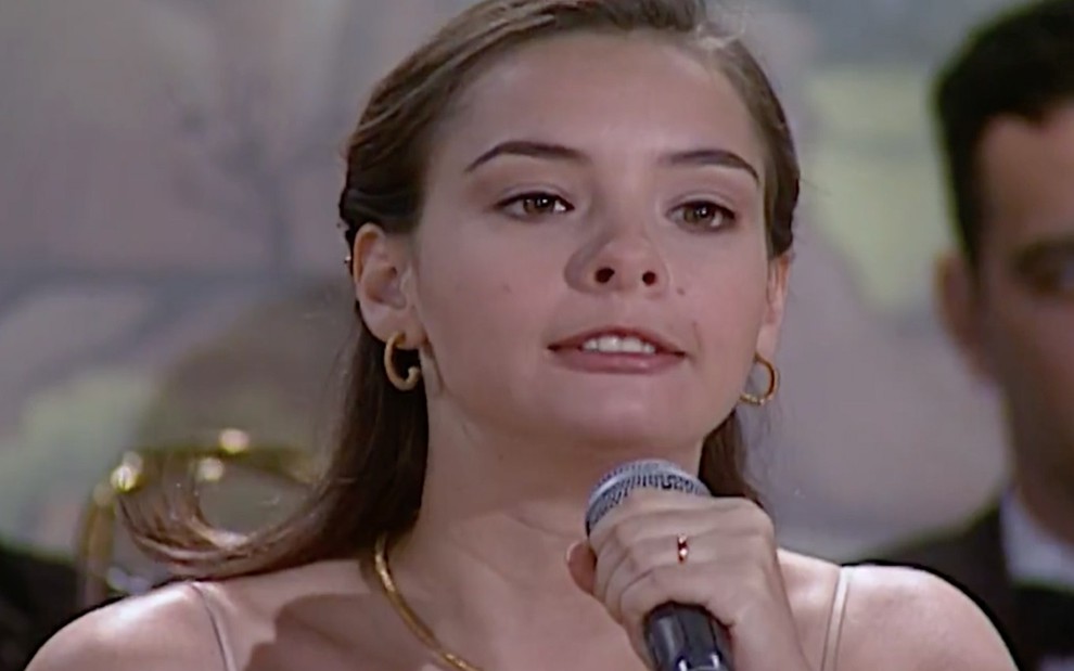 Regiane Alves em cena de Laços de Família: personagem segura um microfone com uma das mãos e olha com indignação para alguém fora do quadro