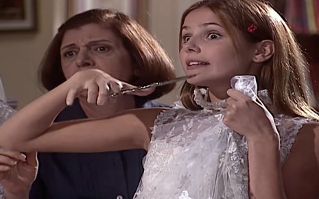 A atriz Deborah Secco como Íris segura uma tesoura com a mão direita e corta um vestido de noiva branco que segura contra o corpo com a mão esquerda em cena de Laços de Família