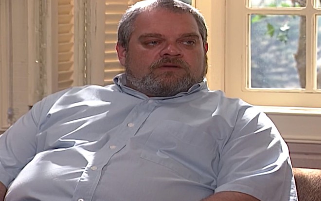 O ator José Victor Castiel, sentado com uma camisa azul, em cena como Viriato em Laços de Família