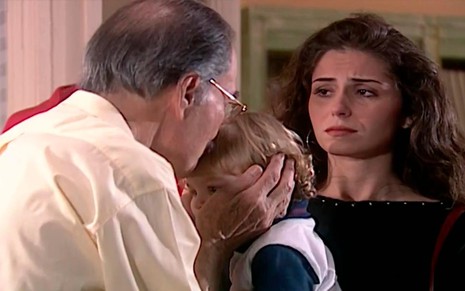 Leonardo Villar e Giovanna Antonelli em cena de Laços de Família: ator está de costas e deixa criança em seu colo, enquanto isso, atriz olha aflita a situação em segundo plano